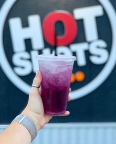 a hand holding a purple drink in front of a hot shots sign