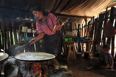 a woman cooking food over an open fire in a hut with other people sitting around