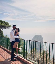 a man and woman posing for a photo on a balcony overlooking the ocean with mountains in the background