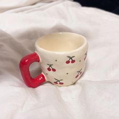 a white cup with cherries painted on it and a red handle sitting on a bed