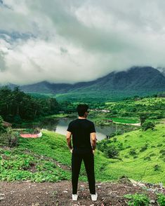 a man standing on top of a lush green hillside next to a lake and mountains