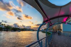 the sun is setting on the water and people are walking down the walkway by the river