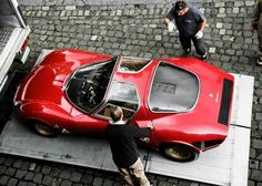 two men standing next to a red car on top of a parking lot with the hood open