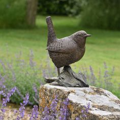 a bird statue sitting on top of a rock in front of purple flowers and trees