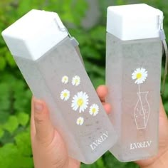 two plastic bottles with daisies on them are held in front of green plants and bushes