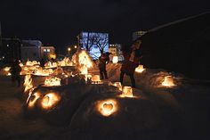 some people are standing in the snow with many lit up candles around them and buildings behind them