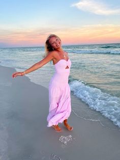 a woman in a pink dress is walking on the beach with her arms spread out