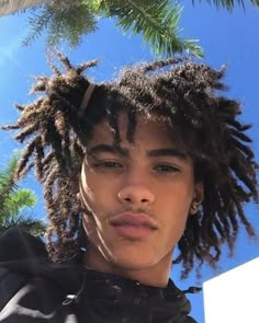 a young man with dreadlocks standing in front of a palm tree and blue sky