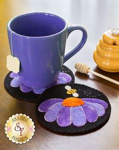 two coasters with purple flowers on them next to a honey comb and a mug