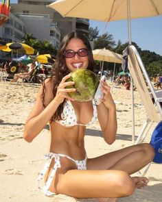 a woman sitting on the beach holding a green fruit in her hand and smiling at the camera