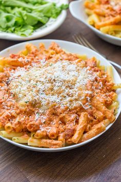 two bowls filled with pasta and sauce on top of a wooden table next to salad