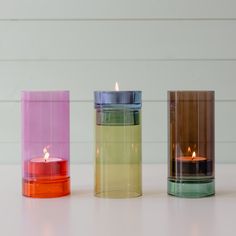 three different colored candles sitting next to each other on a white counter top in front of a wall