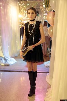 a woman in a black dress and pearls holding a purse standing next to a white curtain