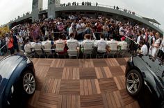 two cars parked in front of a crowd of people on a deck with chairs and tables