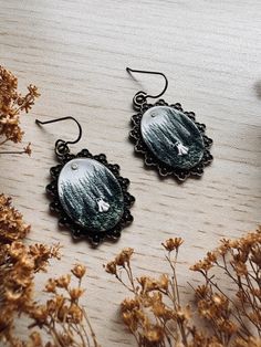 two earrings are sitting on a table next to some dried flowers and plants, one is decorated with an image of a bird