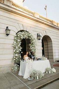 a bride and groom sitting at a table in front of a building with white flowers