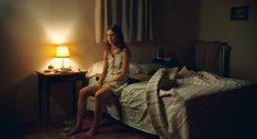 a woman sitting on top of a bed next to a night stand with a lamp