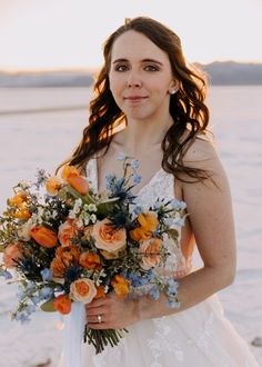 a woman holding a bouquet of flowers in her hands and looking at the camera while standing on a beach