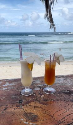 two drinks sitting on top of a wooden table next to the ocean in front of a palm tree