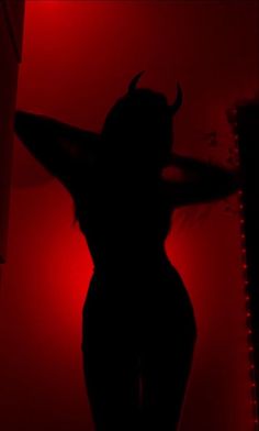 the silhouette of a woman with horns on her head and hands behind her head, in front of a red light