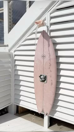 a pink surfboard leaning against the side of a white building with shutters on it