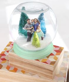 a snow globe with family in it on top of a wooden block next to a christmas tree