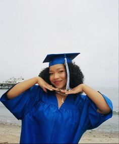 a woman wearing a blue graduation gown and holding her hands to her face while standing in front of the ocean