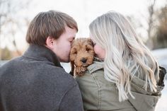 a man and woman kissing while holding a small dog in their lap with snow on the ground behind them