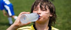 a young boy drinking water from a plastic bottle while standing on a soccer field with other players in the background