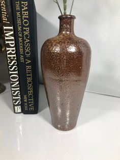 a brown vase sitting next to a book on a white counter top with a flower in it