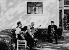 black and white photograph of three men playing instruments in front of a house with bushes