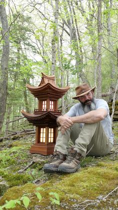 a man sitting on the ground next to a small wooden structure in the woods with trees