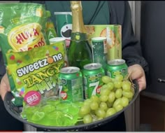 a person holding a tray full of sodas and green grapes in front of them