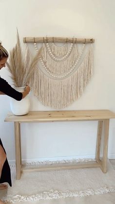 a woman in black dress holding a white vase next to a wooden bench and wall hanging