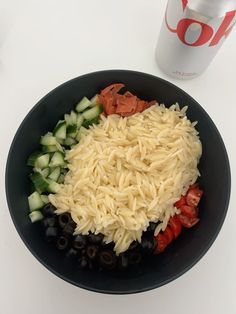 a black bowl filled with pasta and vegetables next to a can of soda on a white table