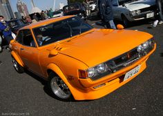 an orange car parked in a parking lot next to other cars and people standing around