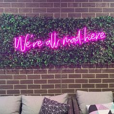 we're all mad here neon sign on brick wall above couches with pillows