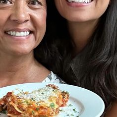 two women are smiling and posing for the camera while holding a plate with lasagna on it