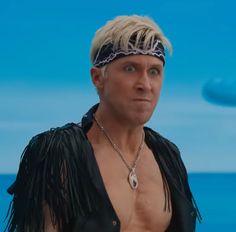 a man with blonde hair and no shirt on standing in front of the ocean looking at something