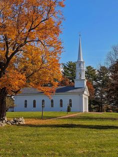 a white church with a steeple surrounded by trees in fall colors and leaves on the ground