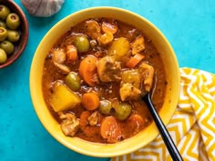 a yellow bowl filled with stew next to an olives and pepper shaker on a blue surface