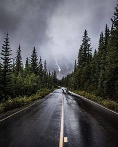 an empty road in the middle of some trees and mountains with snow on it, under a cloudy sky