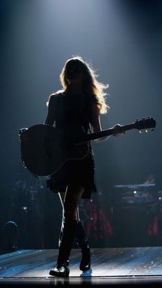 a woman holding a guitar in her right hand and standing on a stage with lights behind her