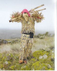 a scarecrow standing on top of a grass covered hill holding two large wooden sticks