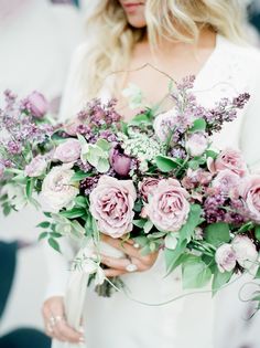 a woman holding a bouquet of flowers with greenery and purple blooms in her hands