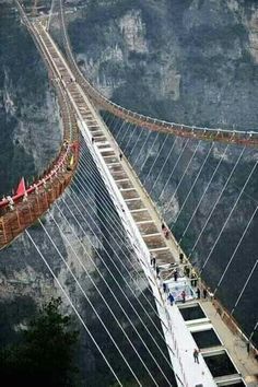 people walking across a suspension bridge in the middle of some mountains, with one person standing on it