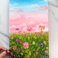 someone is painting flowers in the grass with pink and blue sky behind them on a canvas