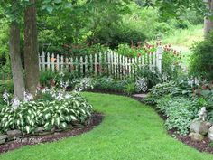 a white picket fence surrounded by lush green grass and flowers in the foreground is a garden with trees, shrubs, and lawn