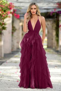 A woman wears a deep burgundy, floor-length dress with layered ruffles, standing confidently in a floral courtyard with soft lighting and greenery in the background, suggesting a romantic setting.