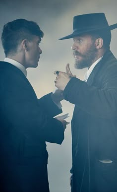 two men standing next to each other in suits and hats, one with his hand on the other's chest